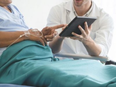 A patient sits up in a hospital bed and points at a tablet being held by a clinician sitting beside them.